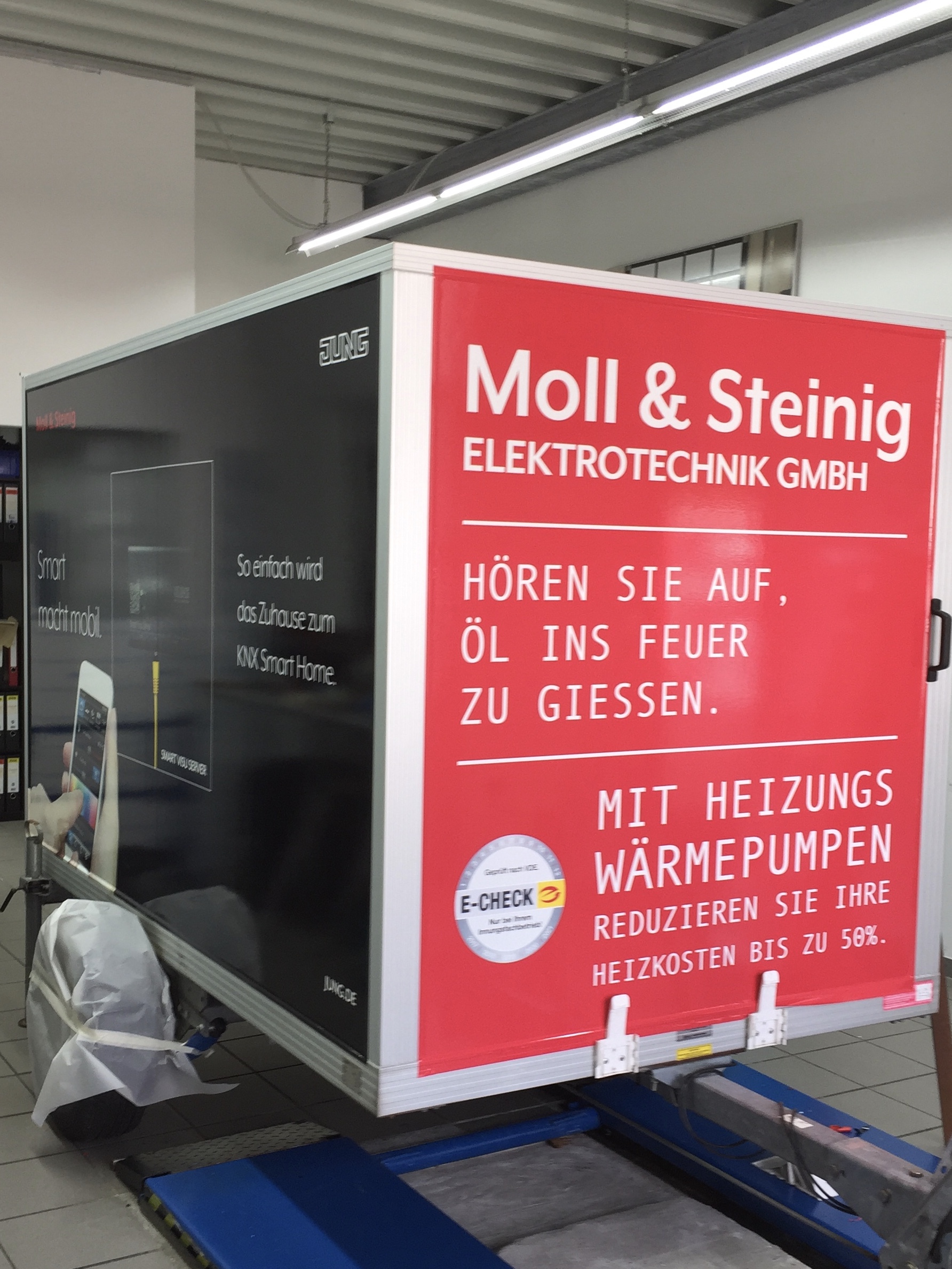 Anhänger der Moll & Steinig Elektrotechnik GmbH mit auffälliger Werbung für Heizungswärmepumpen und E-Check-Service, abgestellt in einer Werkstatt.
