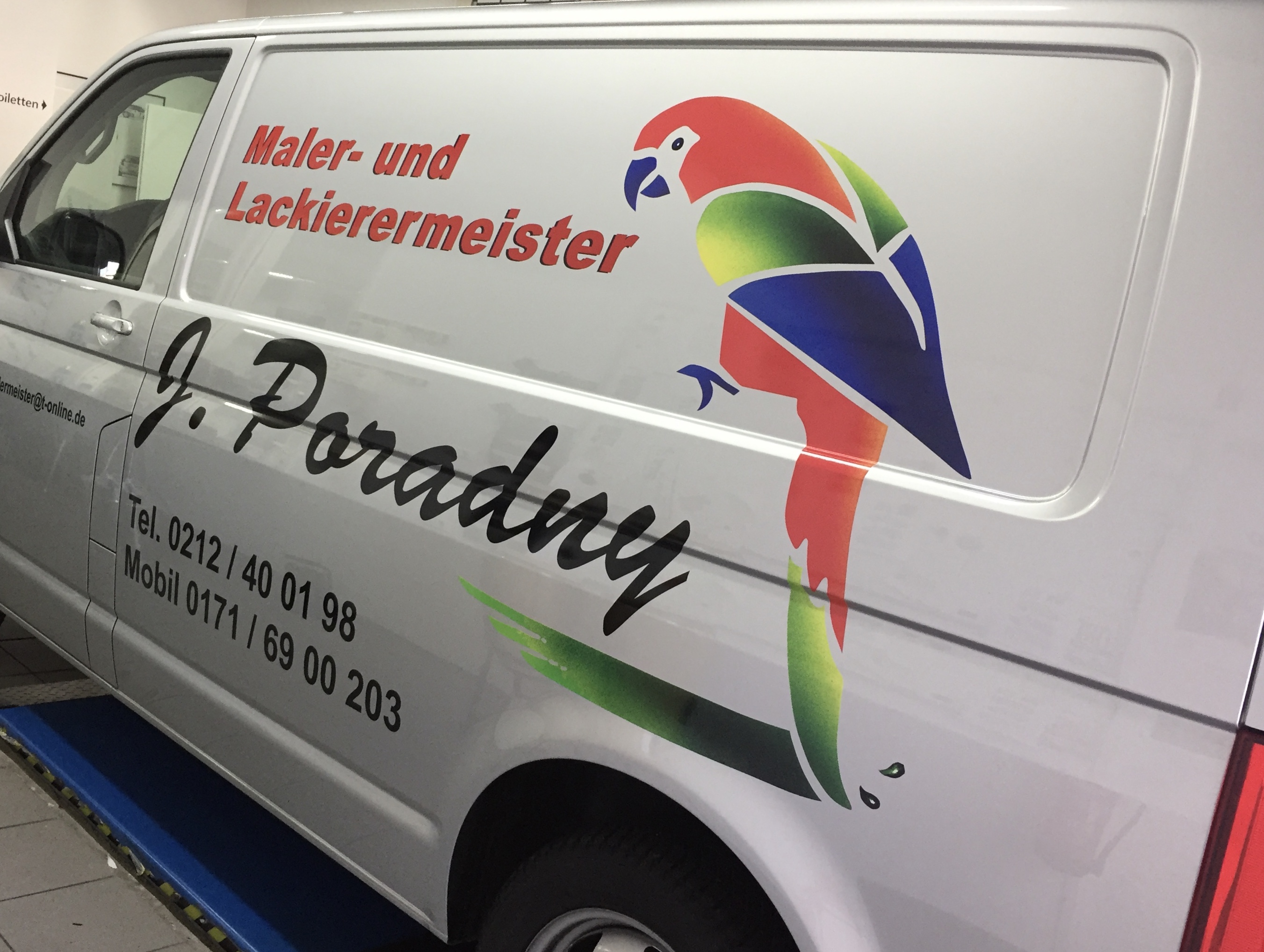 Silberner Transporter mit Logo eines bunten Papageis und Beschriftung 'Maler- und Lackierermeister J. Poradny', abgestellt in einer Werkstatt.