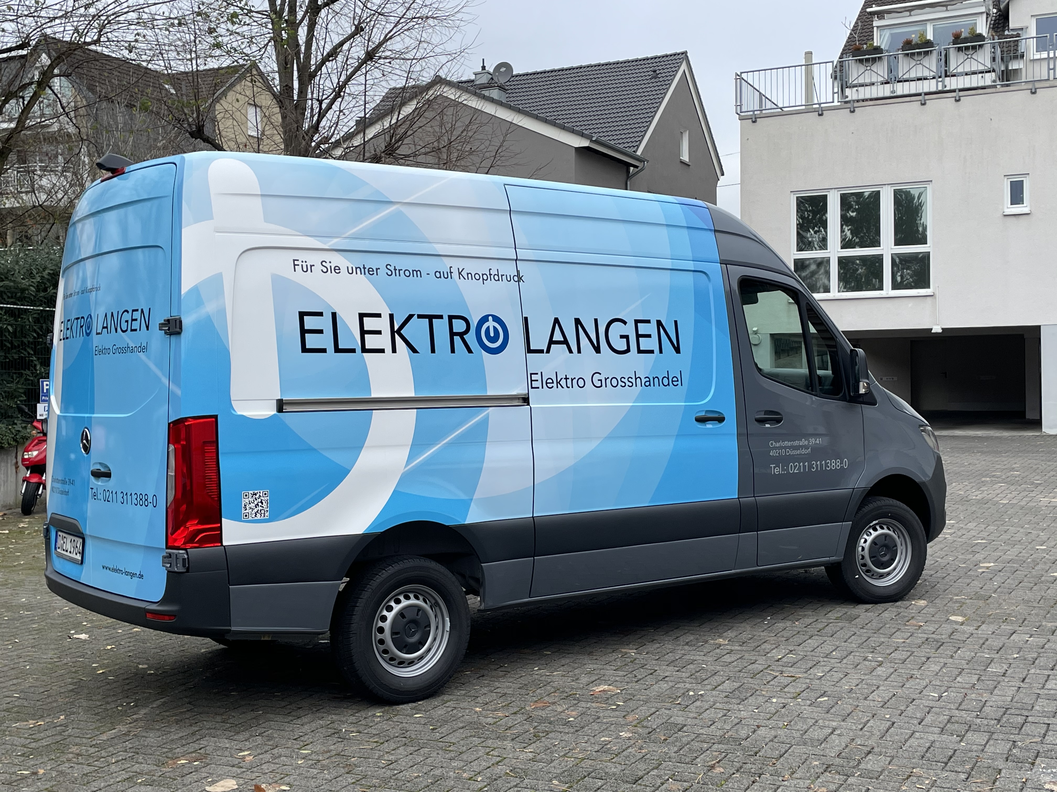 Blau-weißer Elektro Langen Lieferwagen mit Firmennamen und Slogan 'Für Sie unter Strom – auf Knopfdruck', geparkt vor einem Wohngebäude.