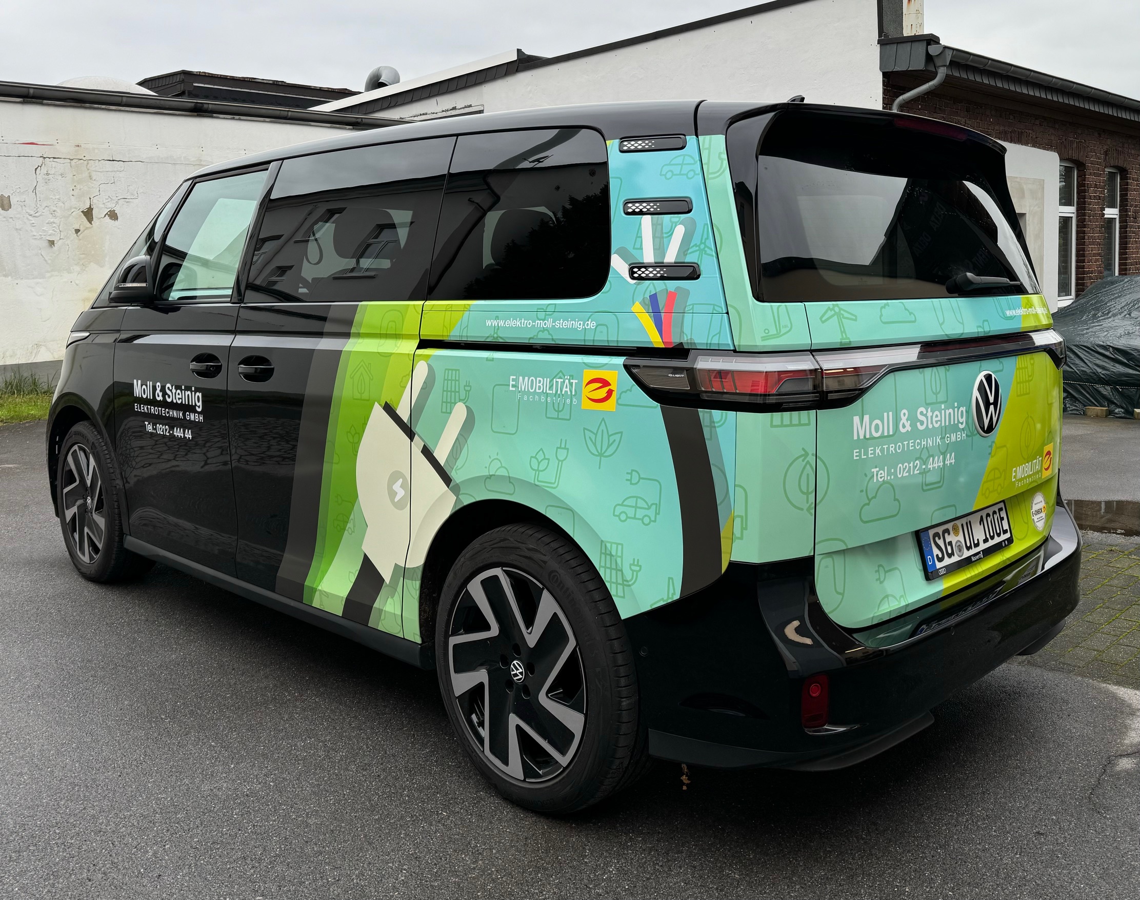 Schwarzer und grüner VW ID.Buzz der Moll & Steinig Elektrotechnik GmbH mit Werbebeklebung zu E-Mobilität, geparkt auf einem Parkplatz.