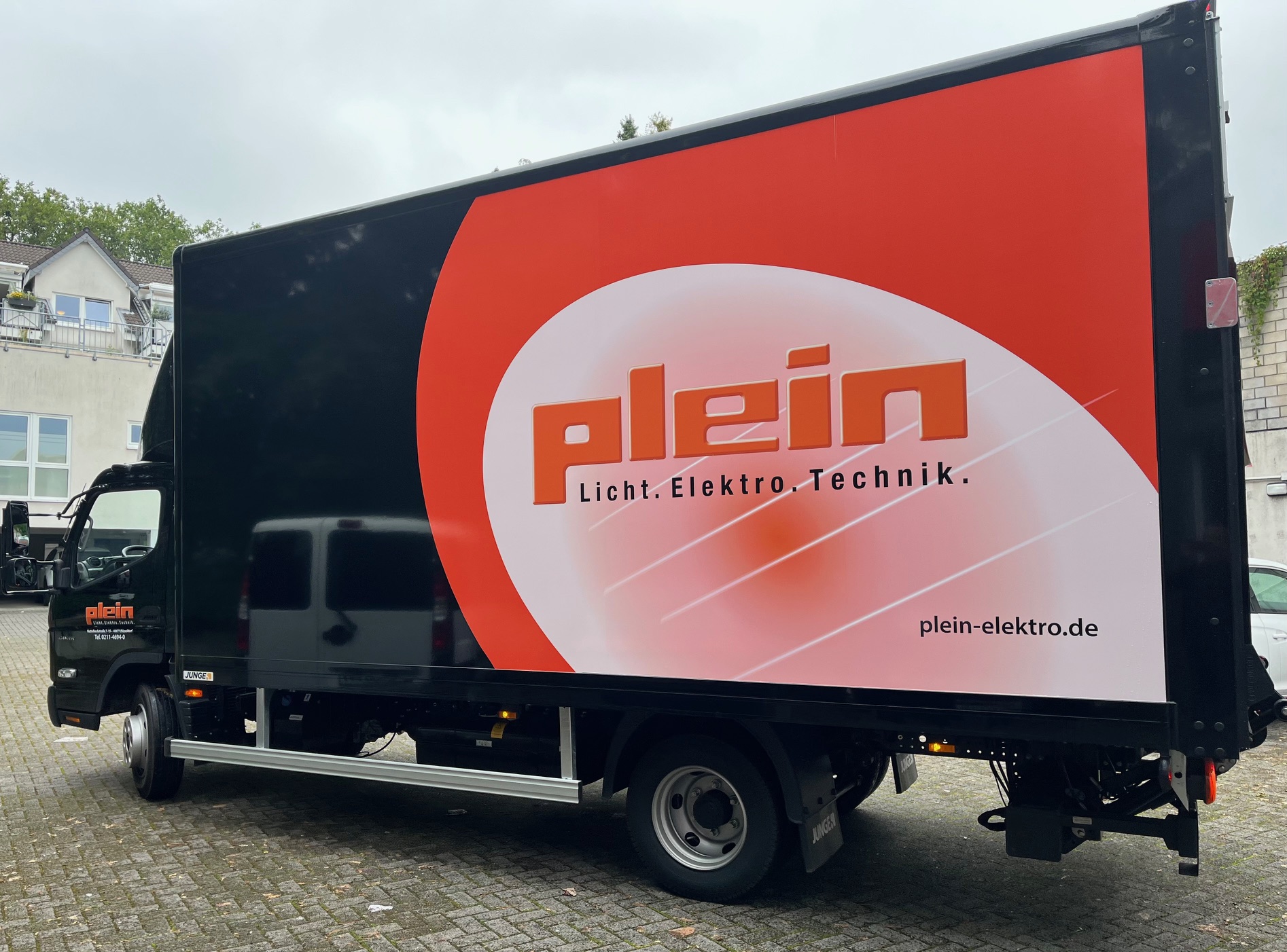 Schwarzer Lastwagen mit großem, rotem 'Plein Licht. Elektro. Technik.'-Logo und Webadresse, geparkt auf einem gepflasterten Hof.