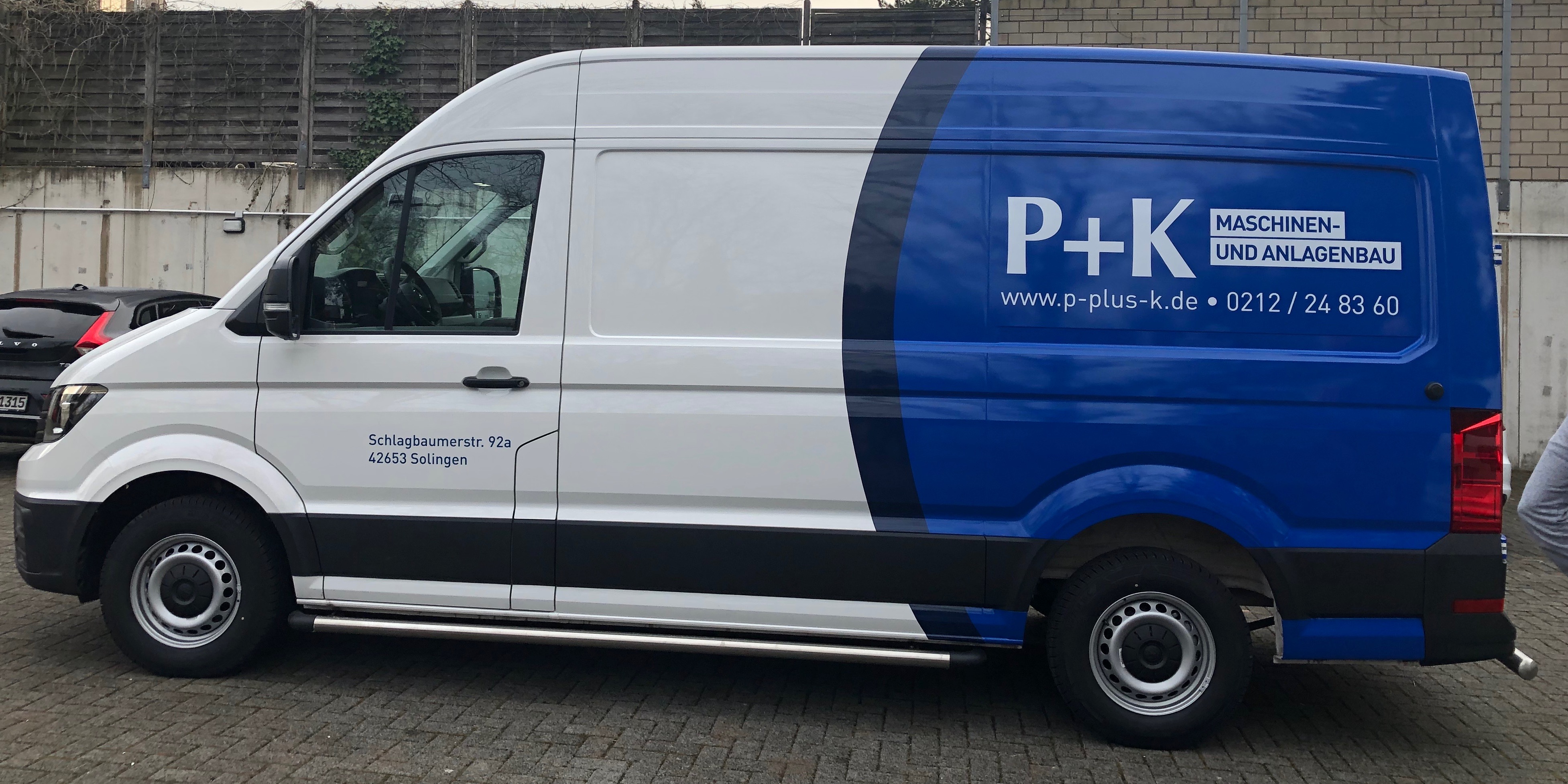 Blau-weißer Transporter der Firma P+K Maschinen- und Anlagenbau mit Firmenanschrift und Webadresse, geparkt vor einer Mauer.