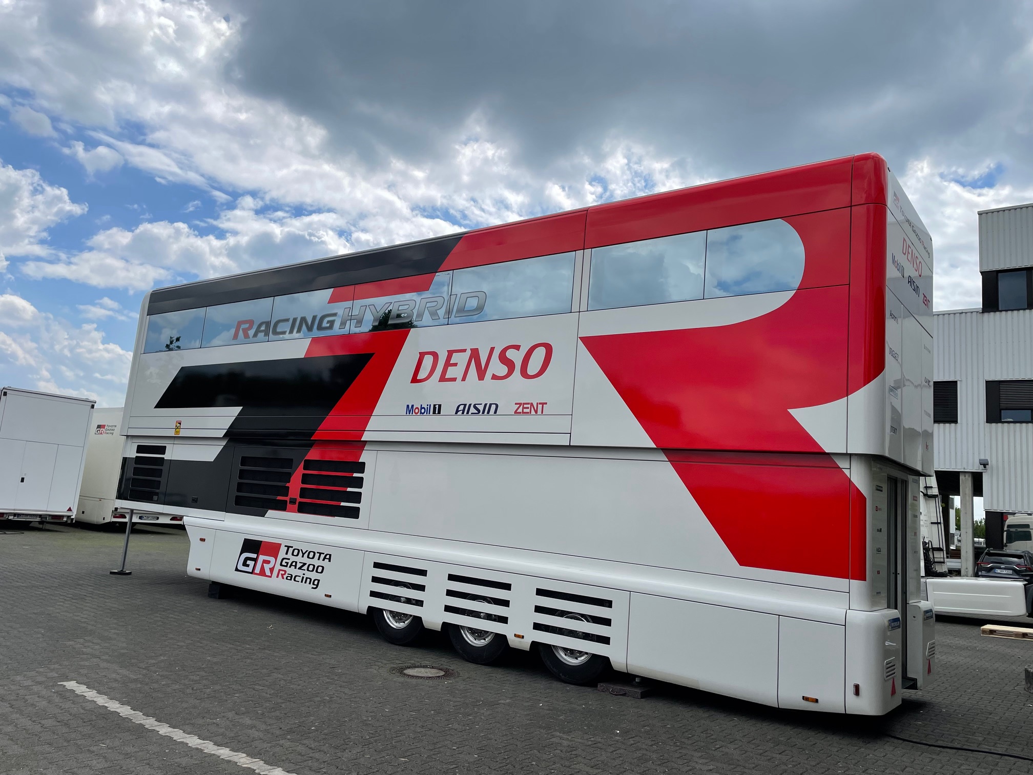 Doppelstöckiger Toyota Gazoo Racing Hybrid-Transporter in auffälligem Rot-Weiß-Design mit Denso- und Mobil1-Branding, geparkt auf einem Industriehof unter bewölktem Himmel.