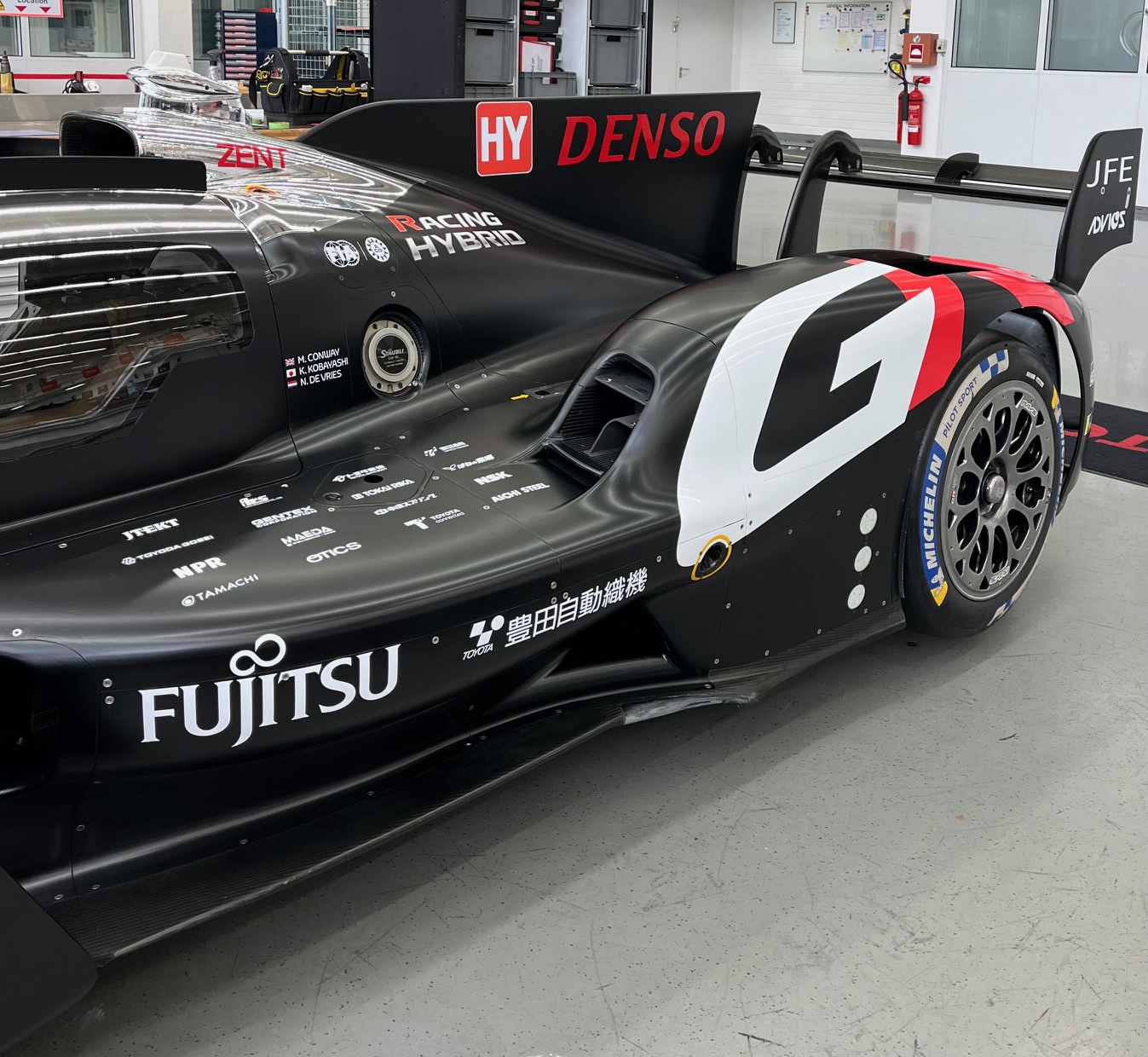 Seitenansicht eines Toyota Racing Hybrid-Rennwagens mit Denso- und Fujitsu-Logos, ausgestattet mit Michelin-Reifen und auffälligem aerodynamischem Design, aufgenommen in einer Werkstattumgebung.